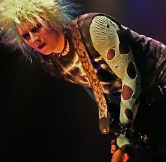 A person with spiky, bleach-blonde hair, wearing a dark eye makeup and a snake-patterned top. The style is punk-inspired, featuring a mesh sleeve with large holes and a belt adorned with chains.