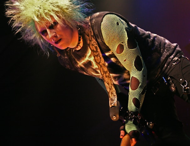 A person with spiky, bleach-blonde hair, wearing a dark eye makeup and a snake-patterned top. The style is punk-inspired, featuring a mesh sleeve with large holes and a belt adorned with chains.