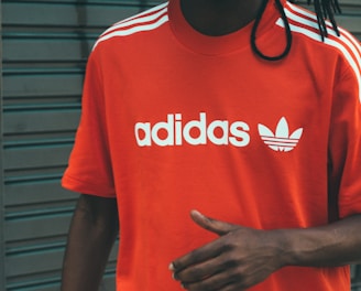A person wearing a bright red Adidas t-shirt featuring white stripes on the shoulders and the brand's logo printed in white on the front. The background consists of a gray, metal roller shutter with some graffiti on the left side.