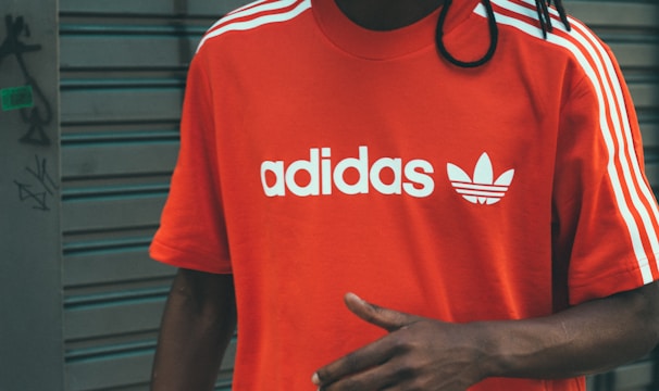 A person wearing a bright red Adidas t-shirt featuring white stripes on the shoulders and the brand's logo printed in white on the front. The background consists of a gray, metal roller shutter with some graffiti on the left side.