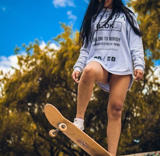 A person stands confidently on a skateboard positioned at the edge of a graffiti-covered ramp, with long hair blowing slightly in the wind. They are wearing a white sweatshirt with black text and graphics, paired with white sneakers. The background features lush green trees under a bright blue sky with scattered clouds.
