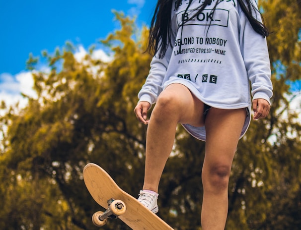 A person stands confidently on a skateboard positioned at the edge of a graffiti-covered ramp, with long hair blowing slightly in the wind. They are wearing a white sweatshirt with black text and graphics, paired with white sneakers. The background features lush green trees under a bright blue sky with scattered clouds.