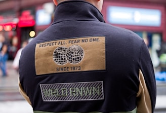 A person wearing a black cap and a color-blocked jacket with patches is standing with their back turned in an urban setting. The jacket has a design with text stating 'Respect All. Fear No One.' along with images of two globes and other text such as 'Since 1973' and 'WRLD RNWN'. The background shows a red-brick building with large windows and some signs indicating a busy street.