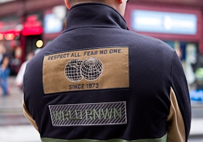 A person wearing a black cap and a color-blocked jacket with patches is standing with their back turned in an urban setting. The jacket has a design with text stating 'Respect All. Fear No One.' along with images of two globes and other text such as 'Since 1973' and 'WRLD RNWN'. The background shows a red-brick building with large windows and some signs indicating a busy street.
