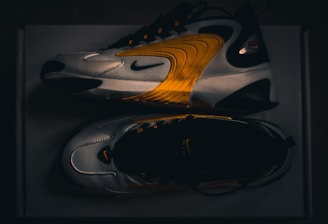 A pair of modern athletic shoes featuring a sleek design with curved lines and a blend of white, black, and vibrant orange hues. The shoes are displayed from a side and top view, highlighting their dynamic style and brand logo.