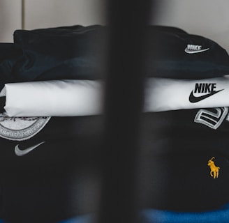 A neatly stacked pile of clothing with visible brand logos, including Nike and a horse logo. The clothes are mainly in dark and neutral colors, with a prominent black piece featuring a large white swoosh. A subtle vertical obstruction divides the image, adding a sense of depth.