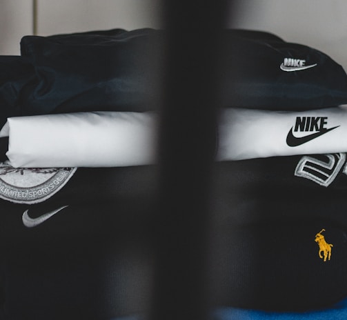A neatly stacked pile of clothing with visible brand logos, including Nike and a horse logo. The clothes are mainly in dark and neutral colors, with a prominent black piece featuring a large white swoosh. A subtle vertical obstruction divides the image, adding a sense of depth.