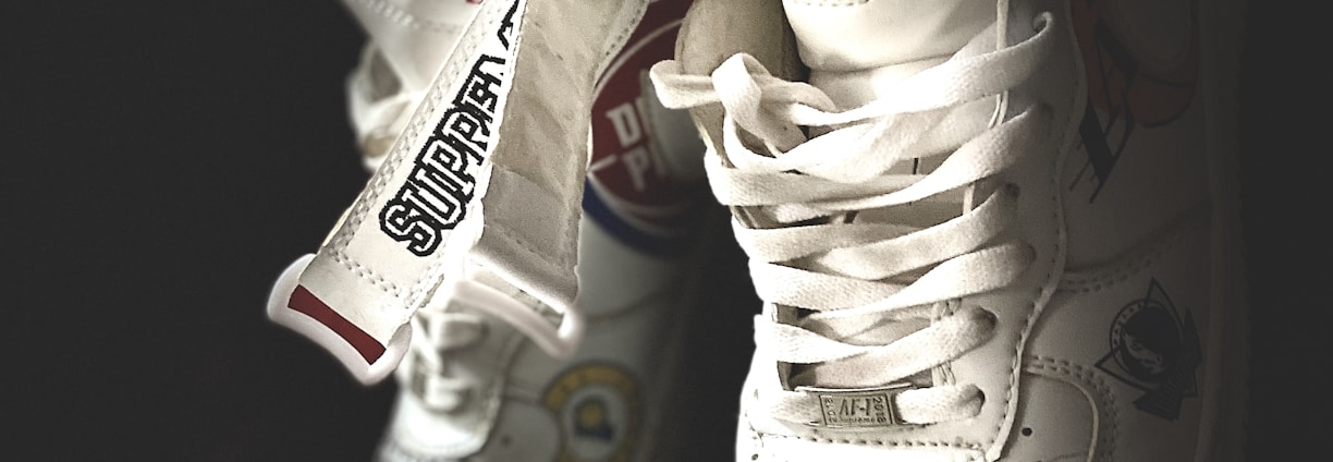 A pair of white sneakers with various logos and text designs, including team names and brand labels. They have a high-top style and intricate detailing on their surface, showcasing a blend of sporty and trendy aesthetics.
