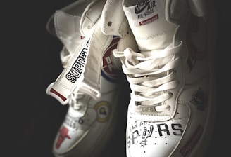 A pair of white sneakers with various logos and text designs, including team names and brand labels. They have a high-top style and intricate detailing on their surface, showcasing a blend of sporty and trendy aesthetics.