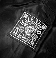 A black jacket features a rectangular patch with white embroidery. The patch includes text and a graphic of an aggressive-looking character with horns. The text on the patch reads 'HALF SUMO', 'FIGHT WEAR', '1985', 'BROOKLYN NY', '20 SAMURAI', and other smaller characters, contributing to a bold and edgy appeal.
