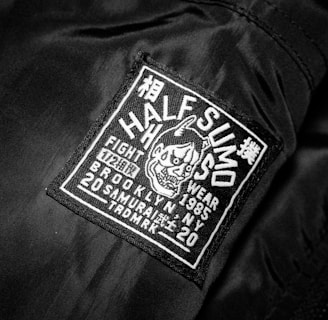 A black jacket features a rectangular patch with white embroidery. The patch includes text and a graphic of an aggressive-looking character with horns. The text on the patch reads 'HALF SUMO', 'FIGHT WEAR', '1985', 'BROOKLYN NY', '20 SAMURAI', and other smaller characters, contributing to a bold and edgy appeal.