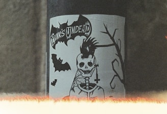 A sticker featuring a skeletal figure with a punk hairstyle and a cross necklace is displayed on a pipe. The text 'Punk's Undead' is visible above the figure. There are bats and a silhouette of a branch in the design. The image has a grainy, vintage look with a light leak effect at the bottom.