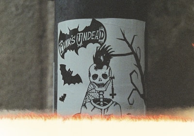 A sticker featuring a skeletal figure with a punk hairstyle and a cross necklace is displayed on a pipe. The text 'Punk's Undead' is visible above the figure. There are bats and a silhouette of a branch in the design. The image has a grainy, vintage look with a light leak effect at the bottom.
