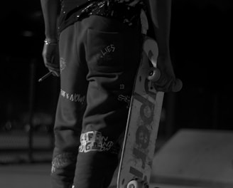 A person dressed in casual, urban attire holds a skateboard under one arm. The image is captured in black and white, emphasizing the high-contrast patterns on the clothing, including a prominent number '23' on the back of the shirt. The setting appears to be an outdoor skate park, indicated by the visible ramps and concrete surface.