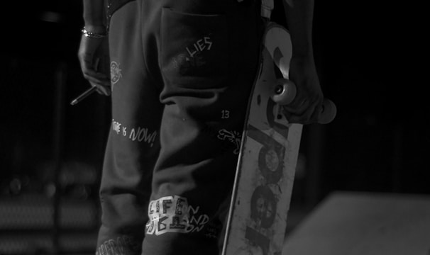 A person dressed in casual, urban attire holds a skateboard under one arm. The image is captured in black and white, emphasizing the high-contrast patterns on the clothing, including a prominent number '23' on the back of the shirt. The setting appears to be an outdoor skate park, indicated by the visible ramps and concrete surface.