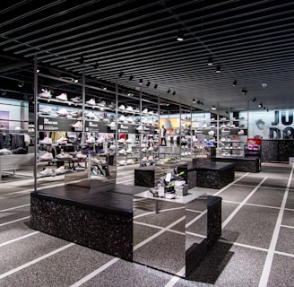 A modern sneaker store with sleek black shelving displaying various athletic shoes and sports apparel. Central seating areas and spotlights provide a contemporary retail ambiance. The signage prominently features a famous sportswear slogan in the background.