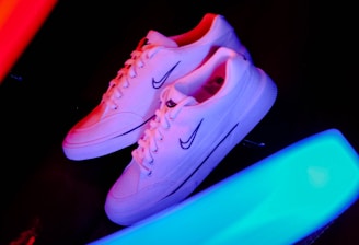 A pair of white sneakers with a prominent logo on the side is placed against a dark background. Bright neon streaks of red and blue light surround the shoes, creating a vibrant and energetic atmosphere.