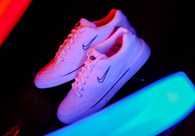 A pair of white sneakers with a prominent logo on the side is placed against a dark background. Bright neon streaks of red and blue light surround the shoes, creating a vibrant and energetic atmosphere.