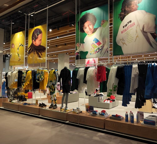A retail store showcases clothing and sportswear on mannequins and hangers, with large vibrant posters featuring children in sports attire above. The interior displays bright garments and accessories on shelves and racks.