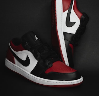 A pair of stylish athletic shoes in black, white, and red colors with distinctive branding. The shoes are positioned against a dark background, showcasing their sleek design and detailed craftsmanship.