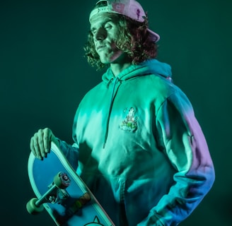 A person wearing a hoodie and a backwards cap holds a skateboard with a cartoon graphic. The lighting is a mix of green and teal, creating a vibrant and edgy vibe.