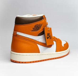 A stylish athletic shoe with a high-top design, featuring a combination of orange, brown, and white colors. The shoe displays a prominent logo on the side and a tag attached to the laces.
