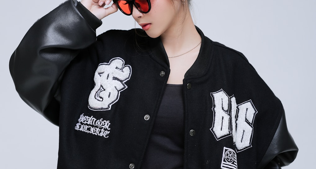 A person wearing a black varsity jacket with embroidered patches, light blue jeans, and a black shirt. They have a checkered headband and red-tinted sunglasses, striking a confident pose against a plain background.