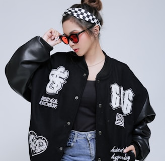 A person wearing a black varsity jacket with embroidered patches, light blue jeans, and a black shirt. They have a checkered headband and red-tinted sunglasses, striking a confident pose against a plain background.