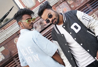 Two individuals are posing in front of a textured metal background, showcasing a casual and stylish demeanor. One is wearing a light blue striped shirt with bold white text, along with vibrant green glasses. The other person sports a black and white varsity jacket layered over a plain white shirt and wears dark sunglasses. Their expressions are confident and relaxed.