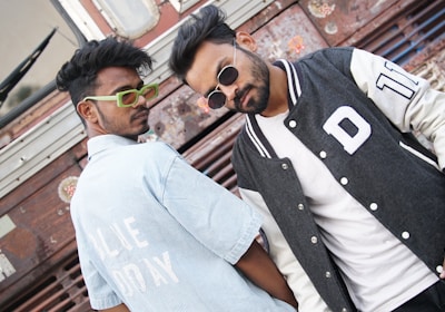 Two individuals are posing in front of a textured metal background, showcasing a casual and stylish demeanor. One is wearing a light blue striped shirt with bold white text, along with vibrant green glasses. The other person sports a black and white varsity jacket layered over a plain white shirt and wears dark sunglasses. Their expressions are confident and relaxed.