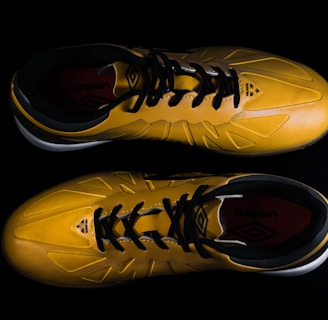 A pair of yellow sports shoes with black laces is placed against a dark background. The design is sleek, with angular patterns on the surface. The inside of the shoes appears to have a red lining with a brand logo visible.