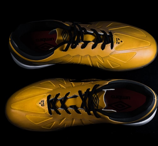A pair of yellow sports shoes with black laces is placed against a dark background. The design is sleek, with angular patterns on the surface. The inside of the shoes appears to have a red lining with a brand logo visible.