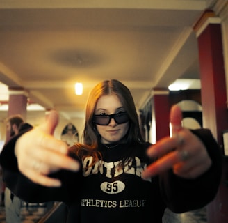 A person with long hair is wearing dark sunglasses and a sweatshirt with the word 'athletics' on it. They have their hands stretched outwards towards the camera, creating a dynamic and energetic pose. The background features an indoor setting with red and white pillars and a patterned floor.
