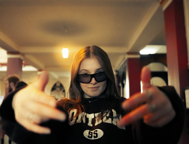 A person with long hair is wearing dark sunglasses and a sweatshirt with the word 'athletics' on it. They have their hands stretched outwards towards the camera, creating a dynamic and energetic pose. The background features an indoor setting with red and white pillars and a patterned floor.