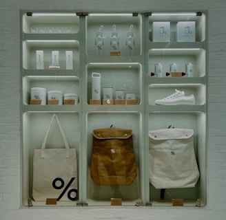 A well-organized display of branded merchandise with a minimalist aesthetic. The items include bottles, cups, a sneaker, bags, and other accessories, all featuring a distinctive percentage symbol on a white or neutral background. The display is set against a white brick wall, giving an industrial feel.