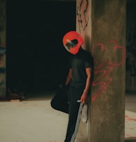 A person wearing a bright red helmet leans casually against a concrete pillar with graffiti on it. The attire is mostly black, featuring a T-shirt and Adidas track pants, complemented by white sneakers. The setting appears to be an unfinished or abandoned industrial space, with dim lighting and a moody atmosphere.