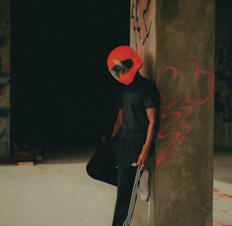 A person wearing a bright red helmet leans casually against a concrete pillar with graffiti on it. The attire is mostly black, featuring a T-shirt and Adidas track pants, complemented by white sneakers. The setting appears to be an unfinished or abandoned industrial space, with dim lighting and a moody atmosphere.