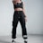 A person stands confidently indoors, wearing black athletic clothing including a crop top and sweatpants. They are also wearing sunglasses, sneakers, and have a watch on their wrist. The stance is assertive, with one hand pointing forward.